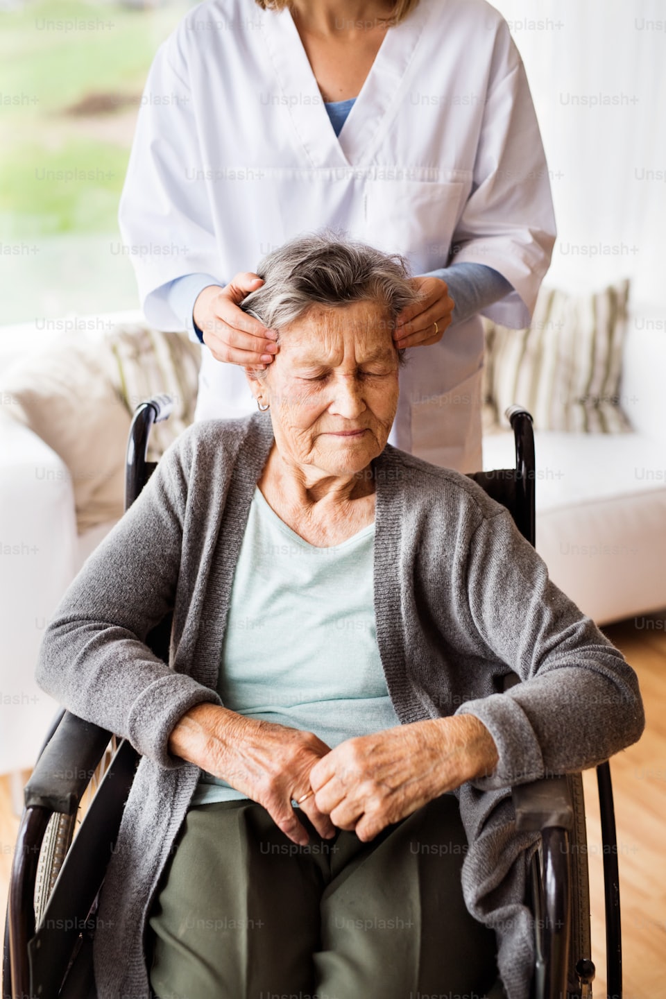 Pflegekraft massiert sanft den Kopf einer Frau im Rollstuhl.